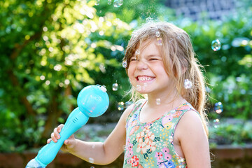 Happy little blonde preschool girl having fun with blowing soap bubble blower. Cute child playing...