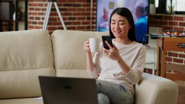 Joyful asian woman texting friend on phone while working remotely on couch in living room. Smiling heartily young adult person texting messages to coworker while doing remote work. Tripod shot
