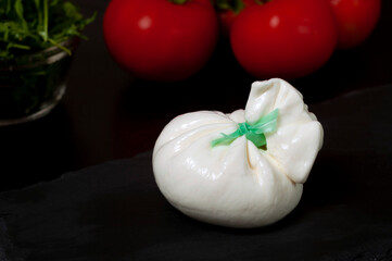 Italian burrata cheese on a black slate board on a background of red tomatoes and spinach and arugula greens.