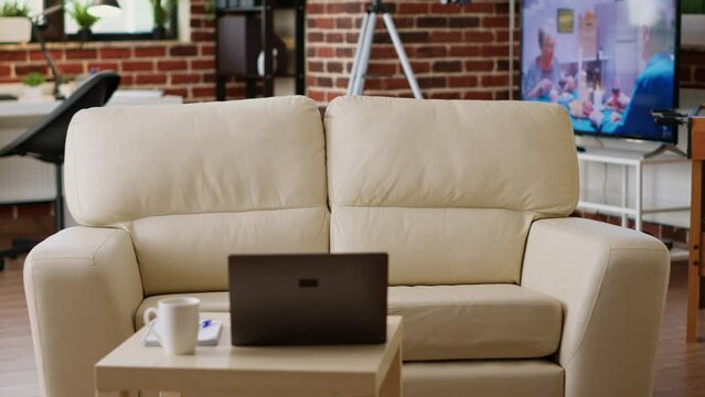 Interior of modern apartment with coffee table having laptop on it. Empty living room with nobody in it and with comfortable sofa and desk having portable computer on top of it.