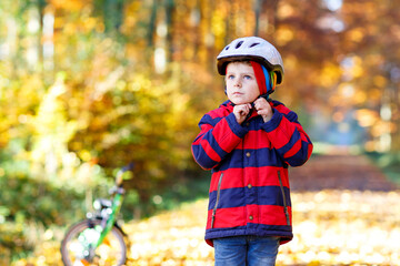 Little kid boy in colorful warm clothes in autumn forest park with a bicycle. Active child putting safe helmet before cycling on sunny fall day in nature. Safety, sports, leisure with kids concept