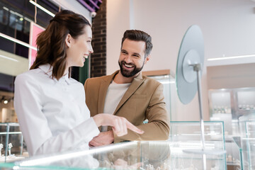 Positive customer looking at young seller pointing at showcase in jewelry shop.