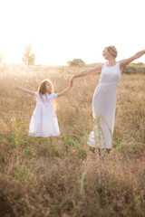 Naklejka na ściany i meble A cute little girl with long blond curly hair and her mother in a white summer dress and a straw boater hat in a field in the countryside in summer at sunset. Nature and Ecolife