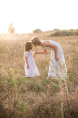 Fototapeta na wymiar A cute little girl with long blond curly hair and her mother in a white summer dress and a straw boater hat in a field in the countryside in summer at sunset. Nature and Ecolife