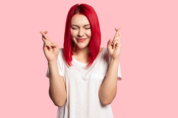 Portrait of lovely girl posing with crossed fingers and waiting for her wish to come true. Adorable model with bright red hairstyle. Happiness and lifestyle concept. Isolated on pink