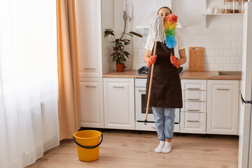 Portrait of young adult Caucasian woman wearing jeans and brown apron, standing in modern kitchen, holding mop and ppduster, washing floor and wiping the dust.