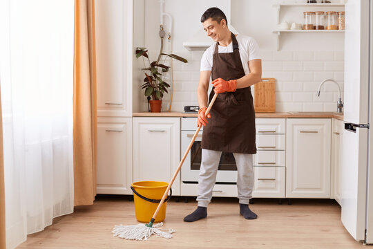 Indoor shot of handsome man brown apron mopping the floor in the kitchen, expressing positive emotions, doing household work, helping his wife with home cleaning.