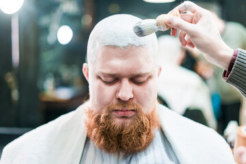 A bearded man is being applied shaving cream on his head.