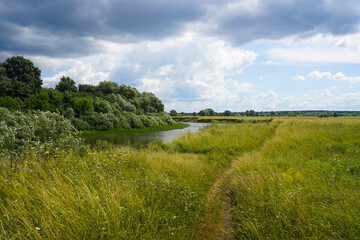 Fototapeta na wymiar Narrow path through meadow by the river and sky with dramatic clouds on summer countryside landscape