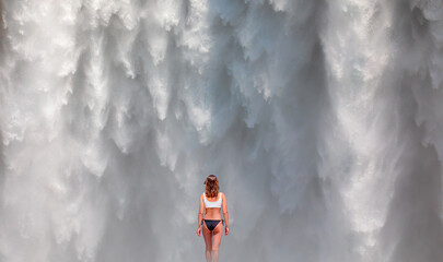 A beautiful girl in a black and white bikini walks towards the waterfall - Icelandic Landscape concept - View of famous Skogafoss waterfall 