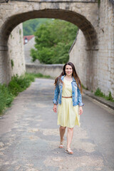Fototapeta premium Lifestyle portrait of young stylish woman with long brunette hair walking on the street in old town, wearing yellow dress and denim jacket