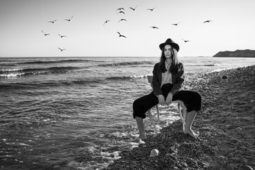 girl in stylish formal suit, shirt and hat sitting on white chair in water of ocean.  Romantic...