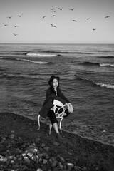 Sea, woman, sky and birds. Moody black and white vertical composition. girl in stylish formal suit, shirt and hat sitting on white chair in water of ocean water. 