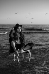 Portrait of beautiful brunette young girl in black suit and hat sitting barefoot on white chair...
