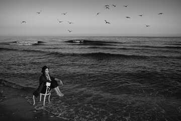 loneliness by the sea. girl in a formal elegant stylish suit, shirt and hat sitting on chair in...