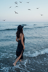 Leaving concept. Romantic beautiful slim girl in long black dress and hat back going away. sea with waves in background. Romantic melancholic composition. Catch me if you can. Blue beautiful sunset