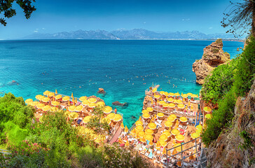 Aerial view of hidden narrow and cozy beach among cliffs and rocks with bright yellow umbrellas and sunbeds and vacationers swimming and chilling in refreshing sea waters