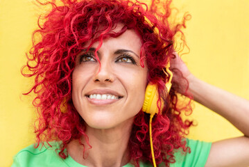 portrait of a smiling latin woman with red afro hair wearing a green t-shirt looking up and listening to music with yellow headphones over a yellow background