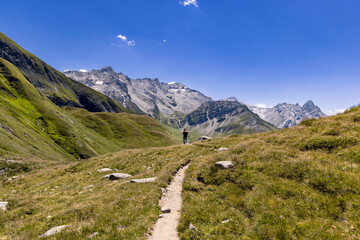 View of a very large and majestic alpine landscape in the Lord of the Rings style. In the foreground a girl walks toward the high mountains in the background, that dominate scene.