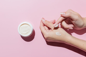Female applying a collagen firming cream to a hand. Open cream jar on pink background. Cosmetics...