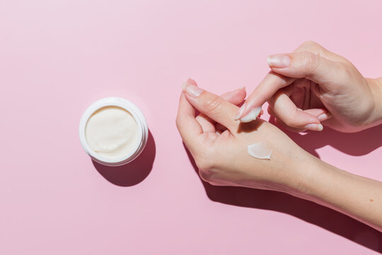 Female applying a collagen firming cream to a hand. Open cream jar on pink background. Cosmetics mockup. Trendy colors and shoot.
