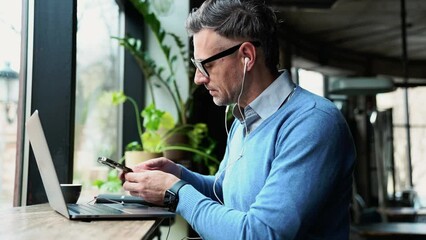 Confident grey haired man working on laptop and looking at phone while sitting in cafe