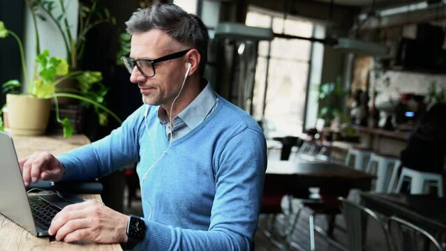 Happy grey haired man working on laptop and drinking coffee while sitting in cafe