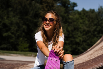 Happy smiling girl with skate board sitting on skate playground and having fun. Extreme sport lifestyle. Laughing child with skate board posing on sport ramp.