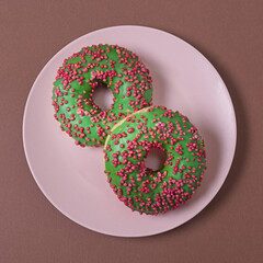 Two green donuts with red and pink sprinkles on a pink saucer on a brown background. View from above