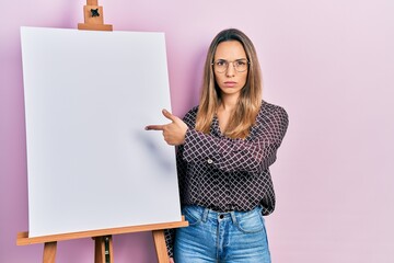 Beautiful hispanic woman pointing to painter easel stand skeptic and nervous, frowning upset...