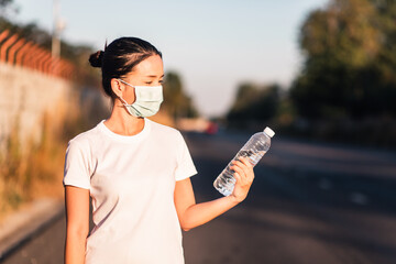 Young Asian woman wearing medical face mask and looking at bottle of clean water in her hand. Coronavirus or covid-19 exercising concept. Beautiful road on blurred background. 