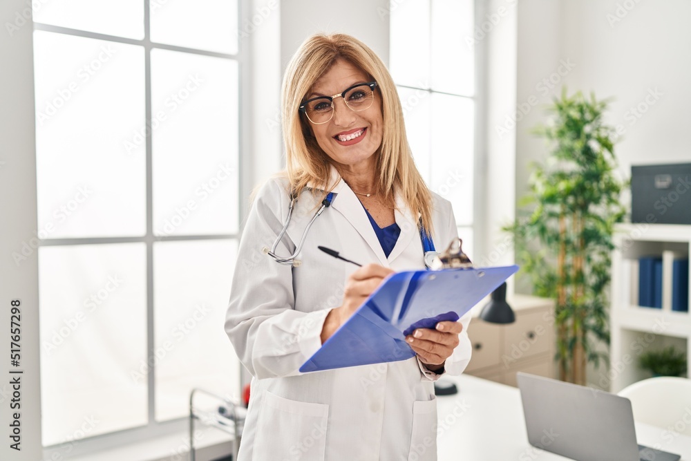 Sticker Middle age blonde woman wearing doctor uniform writing on checklist at clinic