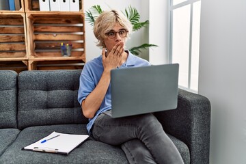 Young man working using computer laptop sitting on the sofa covering mouth with hand, shocked and...