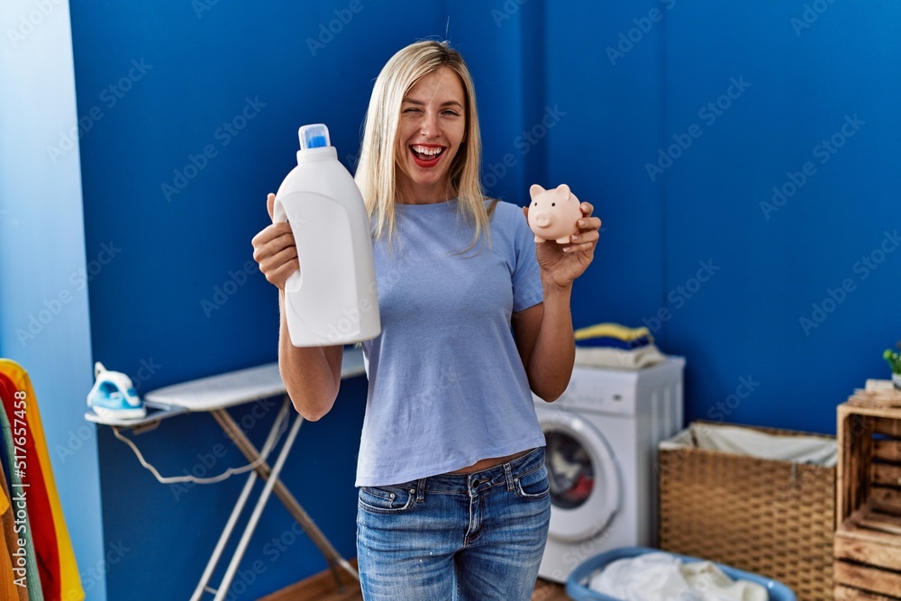 Poster beautiful woman doing laundry holding detergent bottle and piggy bank winking looking at the camera 