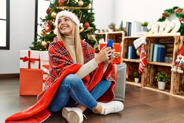 Young blonde woman using smartphone sitting by christmas tree at home