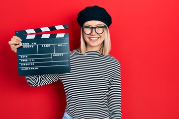 Beautiful blonde woman holding video film clapboard looking positive and happy standing and smiling...