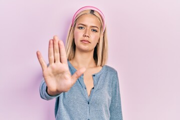 Beautiful blonde woman standing over pink background doing stop sing with palm of the hand. warning...