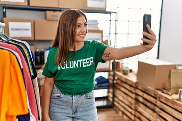 Young hispanic woman wearing volunteer uniform making selfie by the smartphone at charity center