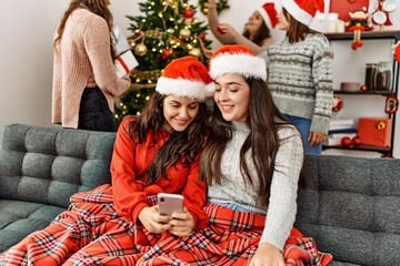 Group of young hispanic women on christmas meeting. Two woman sitting on the sofa using smartphone at home.