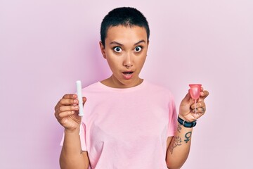 Beautiful hispanic woman with short hair holding menstrual cup and tampon in shock face, looking...