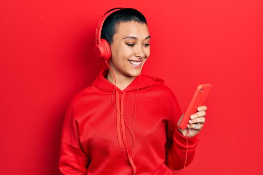 Beautiful hispanic woman with short hair using smartphone wearing headphones looking positive and happy standing and smiling with a confident smile showing teeth