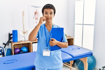 Young hispanic woman with short hair working at pain recovery clinic mouth and lips shut as zip...