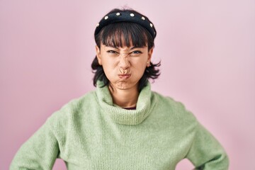Young beautiful woman standing over pink background puffing cheeks with funny face. mouth inflated with air, crazy expression.