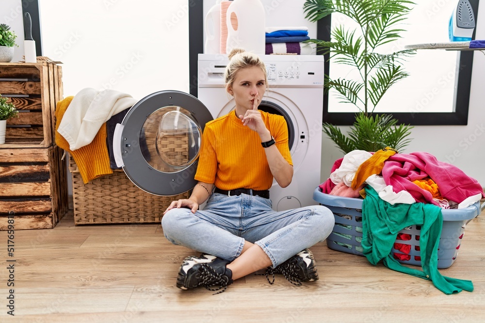 Sticker Young blonde woman doing laundry sitting by washing machine asking to be quiet with finger on lips. silence and secret concept.