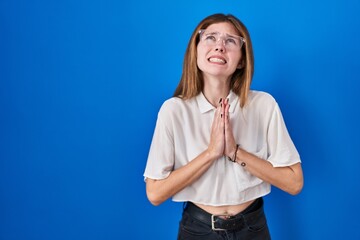 Beautiful woman standing over blue background begging and praying with hands together with hope...