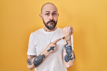 Young hispanic man with tattoos standing over yellow background in hurry pointing to watch time,...