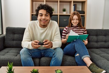 Man playing video game while girlfriend reading book at home.