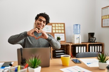 Young hispanic man wearing business style sitting on desk at office smiling in love doing heart symbol shape with hands. romantic concept.