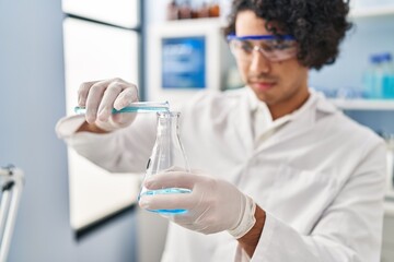 Fototapeta premium Young hispanic man wearing scientist uniform pouring liquid on test tube at laboratory