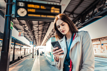 Young woman with departure times behind her waiting for her train while holding her mobile phone -...